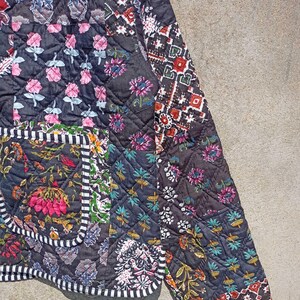 May include: A black quilted jacket with a patchwork design featuring various floral and geometric patterns. The jacket has a white and black trim around the edges and a pocket on the left side.
