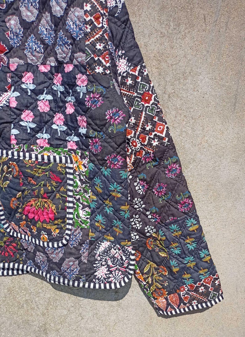 May include: A black quilted jacket with a patchwork design featuring various floral and geometric patterns. The jacket has a white and black trim around the edges and a pocket on the left side.