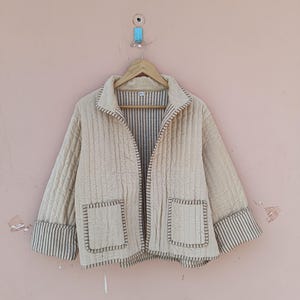 Op de afbeelding: Beige gewatteerde jas met kraag en twee voorzakken. De jas heeft verticale stiksels en gestreepte manchetten. Hij hangt aan een houten hanger tegen een lichtroze muur.