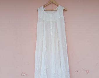 Peut inclure: Une chemise de nuit blanche sans manches suspendue à un cintre en bois. La robe a une encolure ronde, un devant boutonné et une jupe longue et fluide. Le tissu semble léger et légèrement transparent.