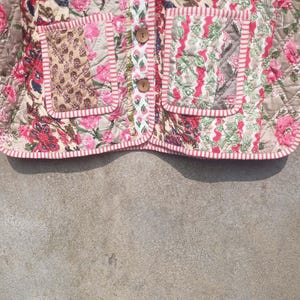 May include: A pink and white quilted jacket with a floral print and a striped trim. The jacket has two pockets on the front and a button closure.