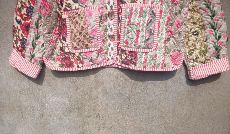 May include: A pink and white quilted jacket with a floral print and a striped trim. The jacket has two pockets on the front and a button closure.
