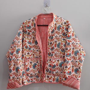 May include: A white quilted jacket with a red and white striped lining. The jacket has a floral print with blue, red, and gold accents. It features two pockets on the front and a button closure.
