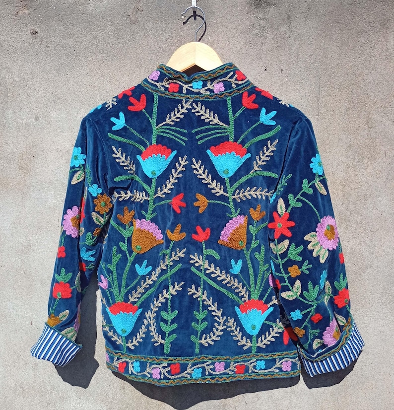 Op de afbeelding: Een blauw fluwelen jasje met kleurrijke bloemenborduurwerk. Het jasje heeft een mandarijnsluiting en lange mouwen.