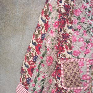 May include: A quilted jacket with a floral print in shades of pink, red, and brown. The jacket has a patch pocket on the front and a striped trim around the edges.
