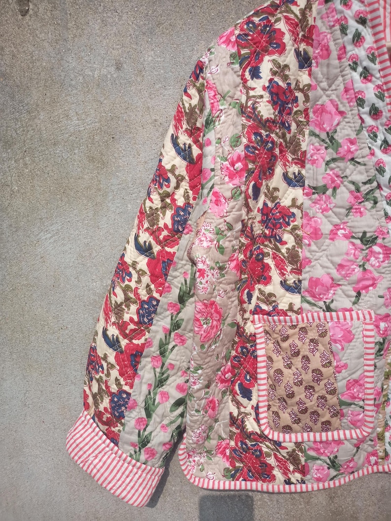 May include: A quilted jacket with a floral print in shades of pink, red, and brown. The jacket has a patch pocket on the front and a striped trim around the edges.