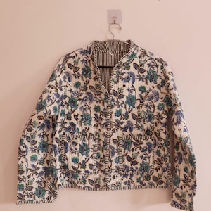 May include: A white quilted jacket with a floral print in shades of blue and green. The jacket has a mandarin collar, a button closure, and two patch pockets.