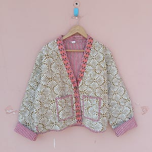 May include: A cream-colored jacket with a brown and white fan-like pattern. The jacket has pink striped trim along the collar, pockets, and cuffs. It hangs on a wooden hanger against a pink wall.