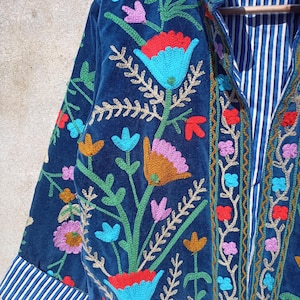 Op de afbeelding: Een donkerblauw fluwelen jasje met kleurrijke bloemenborduursel. Het jasje heeft een mandarijnkraag en lange mouwen met gestreepte manchetten.