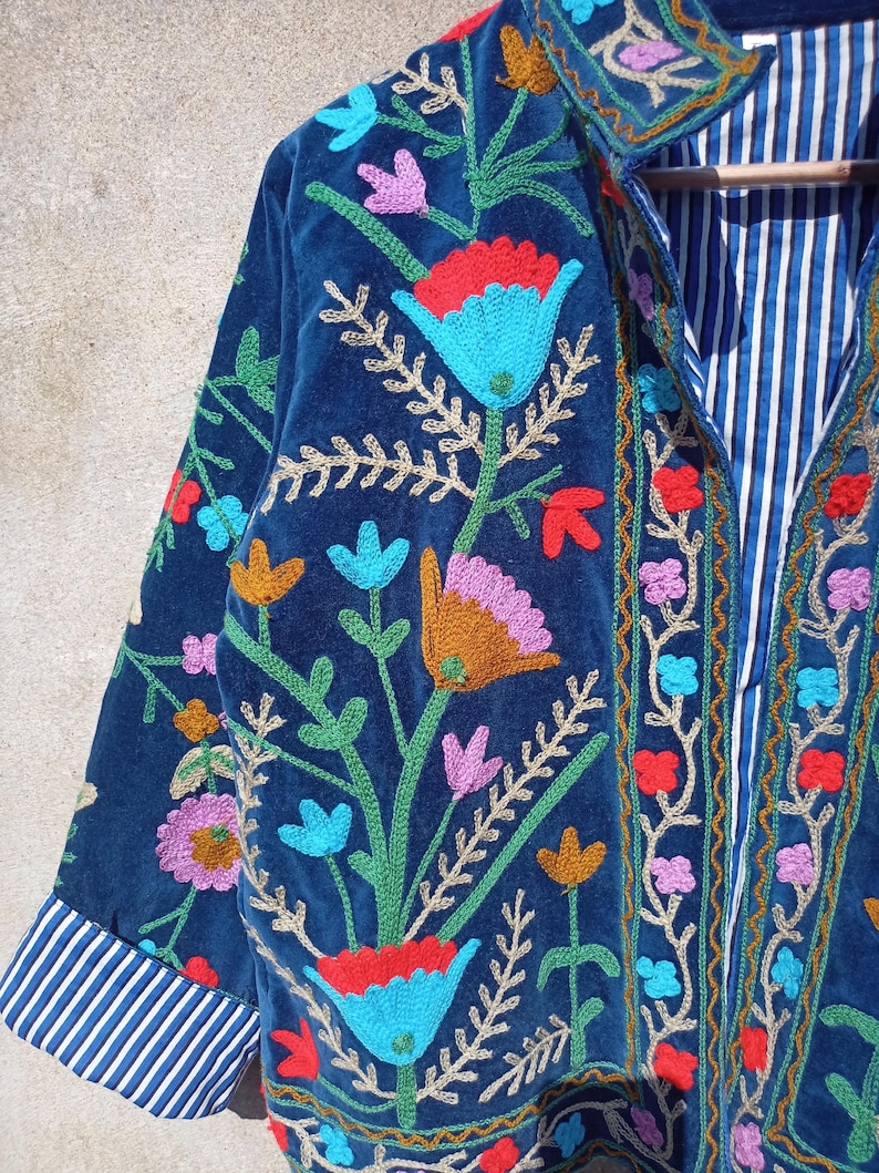 Op de afbeelding: Een donkerblauw fluwelen jasje met kleurrijke bloemenborduursel. Het jasje heeft een mandarijnkraag en lange mouwen met gestreepte manchetten.