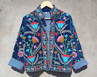Puede incluir: Una chaqueta de terciopelo azul marino con bordado floral de colores. La chaqueta tiene un cierre de botones en la parte delantera y mangas largas con puños a rayas.