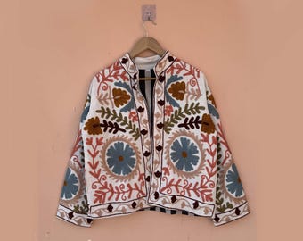 May include: White embroidered jacket with a mandarin collar. The jacket features a floral design in shades of blue, brown, and red. The jacket is buttoned up and hanging on a wooden hanger against a peach-colored wall.