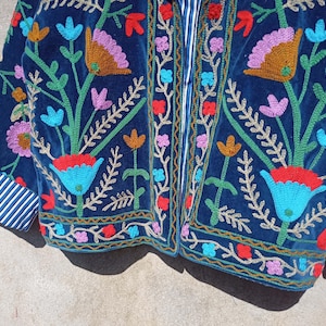 Op de afbeelding: Een blauw fluwelen jasje met kleurrijke bloemenborduursels. Het jasje heeft een ritssluiting en een gestreepte voering aan de manchetten.