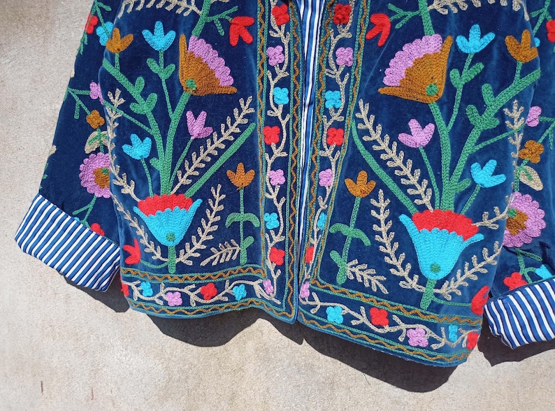 Op de afbeelding: Een blauw fluwelen jasje met kleurrijke bloemenborduursels. Het jasje heeft een ritssluiting en een gestreepte voering aan de manchetten.
