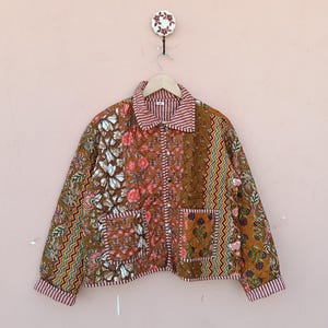 May include: A patchwork jacket with a brown, red, and white floral pattern. The jacket has a collared neckline and two front pockets. The cuffs and collar are striped. The jacket is hanging on a wooden hanger against a pink wall.