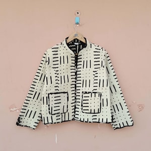 May include: Cream-colored jacket with a black geometric pattern. The jacket has a stand-up collar, two front pockets, and a button-up closure. The jacket is hanging on a wooden hanger against a pink background.