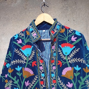Op de afbeelding: Een blauw fluwelen jasje met kleurrijke bloemenborduursels. Het jasje heeft een knoopsluiting aan de voorkant en een kraag.