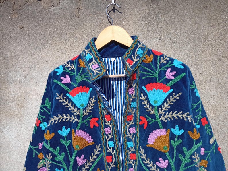 Op de afbeelding: Een blauw fluwelen jasje met kleurrijke bloemenborduursels. Het jasje heeft een knoopsluiting aan de voorkant en een kraag.