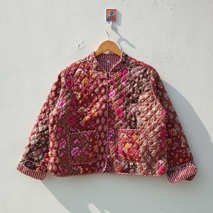 May include: A red quilted jacket with a floral print. The jacket has a button closure and a pocket on the front.