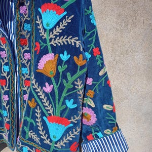 Op de afbeelding: Een blauw fluwelen jasje met kleurrijke bloemenborduursels. Het jasje heeft een mandarijnsluiting en lange mouwen met gestreepte manchetten.