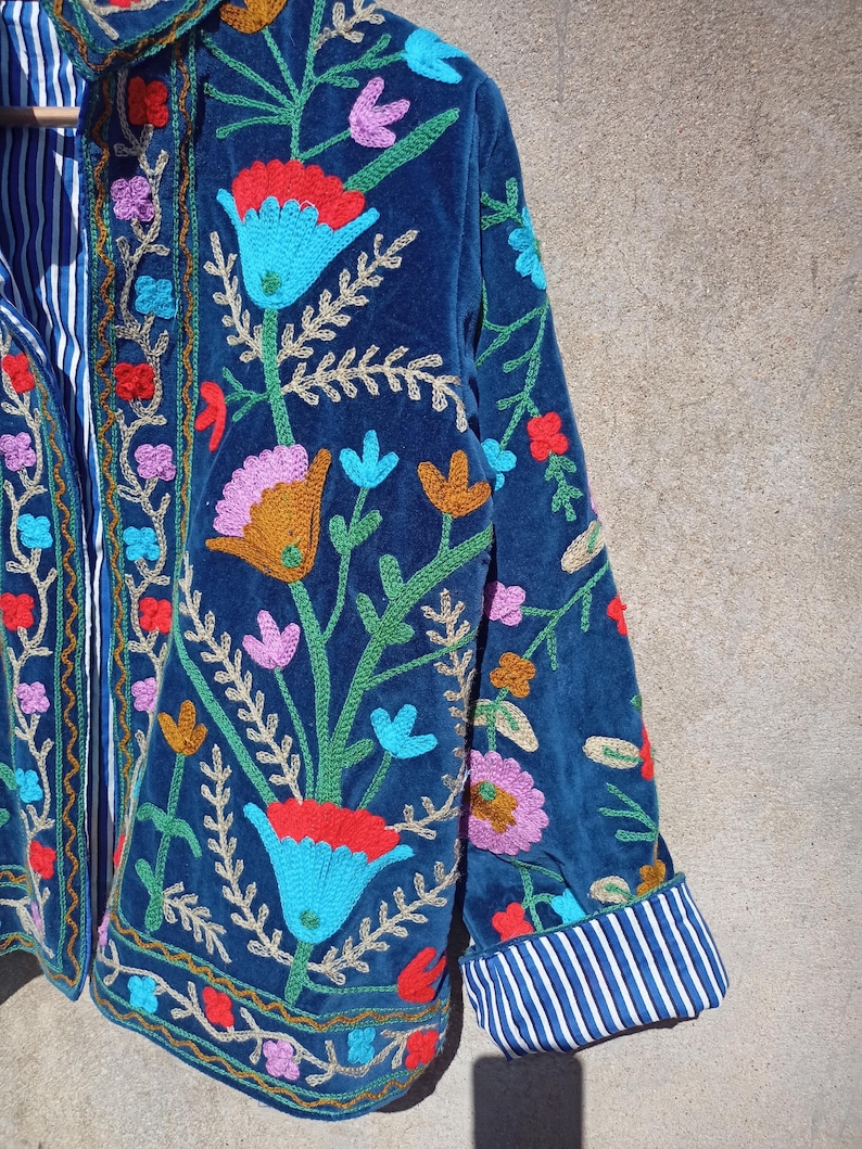 Op de afbeelding: Een blauw fluwelen jasje met kleurrijke bloemenborduursels. Het jasje heeft een mandarijnsluiting en lange mouwen met gestreepte manchetten.