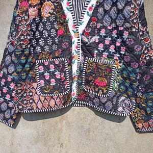 May include: A black quilted jacket with a patchwork design featuring floral and geometric patterns. The jacket has a button closure, two pockets, and a white and black striped trim.