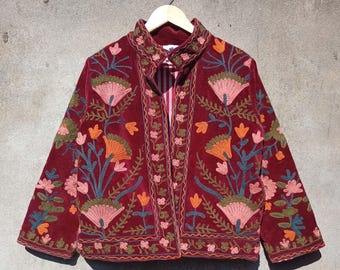 Puede incluir: Una chaqueta de terciopelo burdeos con bordado floral de colores. La chaqueta tiene un cuello mandarín y una cremallera frontal.