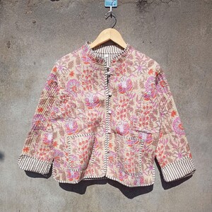 Pode incluir: Uma jaqueta acolchoada bege com estampa floral em tons de rosa, laranja e marrom. A jaqueta tem gola mandarim, fecho de botão e dois bolsos de remendo. As mangas têm uma bainha listrada.