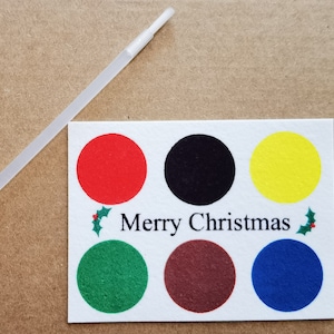 Könnte beinhalten: Eine weiße Karte mit sechs farbigen Kreisen: rot, schwarz, gelb, grün, braun und blau. Die Worte "Merry Christmas" sind darunter gedruckt. Ein klarer Plastikstab befindet sich darüber.