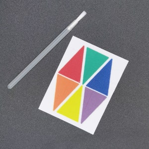 Può includere: Un pennello bianco e un cartoncino bianco con un disegno geometrico arcobaleno. Il disegno è composto da sei triangoli, ognuno di un colore diverso: rosso, arancione, giallo, verde, blu e viola.