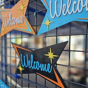 May include: A collection of wooden welcome signs with a retro design. The signs are painted in various colors, including blue, orange, and black. Each sign features a starburst design and the word "Welcome" in a stylized font.