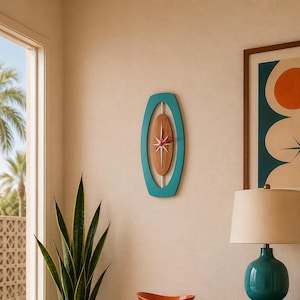 May include: A wooden sideboard with an orange cabinet door and two drawers, topped with a teal lamp and two books titled Palm Springs and Mid-Century Modern. A teal wall clock, an orange bowl, and a snake plant in a speckled pot are also present.