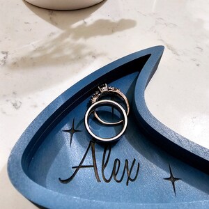 Op de afbeelding: Een blauwe, halvemaanvormige houten ringenschotel met de naam "Alex" erop gegraveerd. Twee zilveren ringen liggen in de schotel.