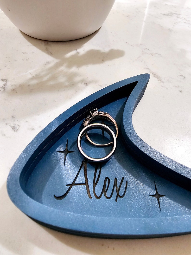 Op de afbeelding: Een blauwe, halvemaanvormige houten ringenschotel met de naam "Alex" erop gegraveerd. Twee zilveren ringen liggen in de schotel.
