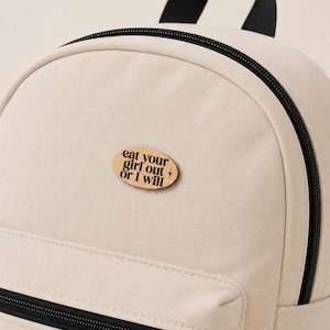 May include: A cream-colored canvas backpack featuring a black zipper closure and a top handle. An oval-shaped wooden patch is attached to the front, displaying the black text "eat your girl out + or I will" in a stylized, bold serif font.