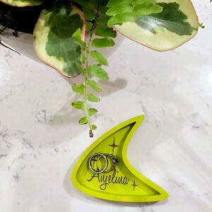 Op de afbeelding: Een felgroene keramische ringenschotel in de vorm van een boemerang met de naam "Ayelina" erop gegraveerd. Twee zilveren ringen liggen in de schotel.
