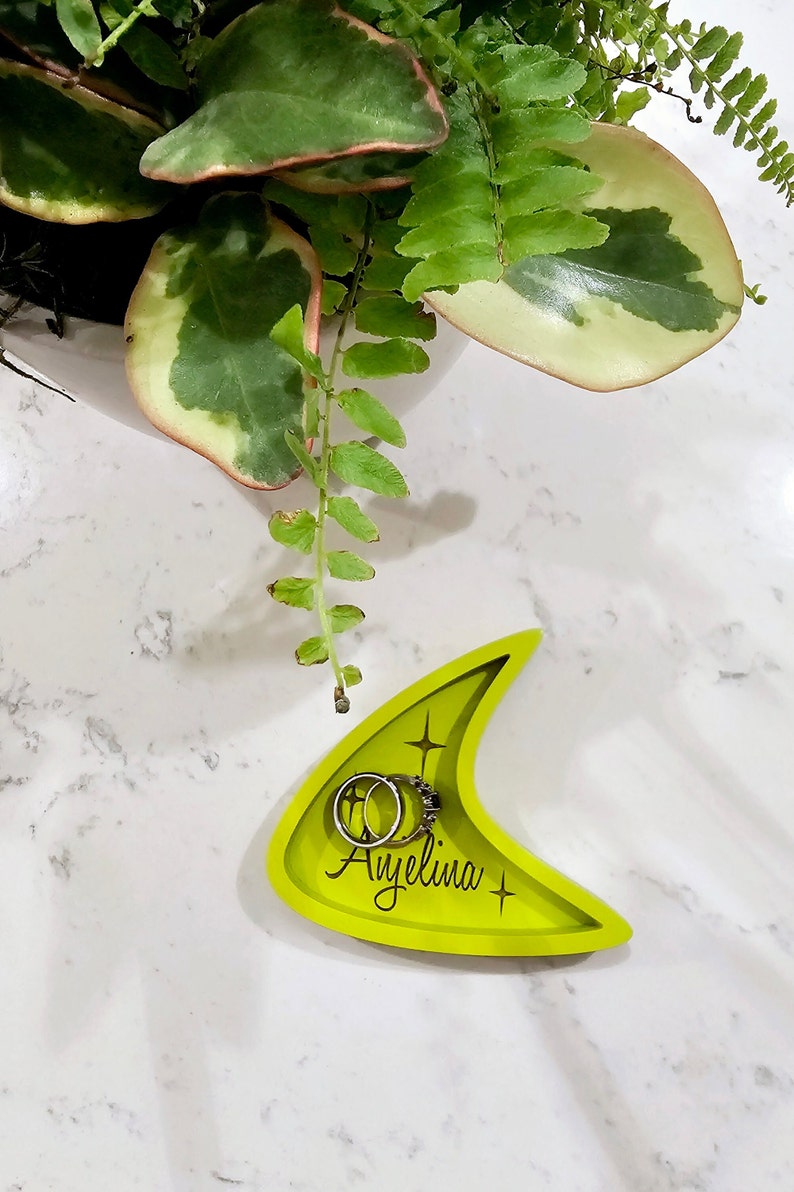 Op de afbeelding: Een felgroene keramische ringenschotel in de vorm van een boemerang met de naam "Ayelina" erop gegraveerd. Twee zilveren ringen liggen in de schotel.