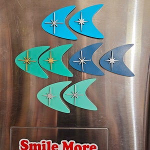 May include: Six retro-style boomerang-shaped magnets with a starburst design in various colors, including teal, turquoise, and green. The magnets are attached to a stainless steel refrigerator with a red "Smile More" logo.