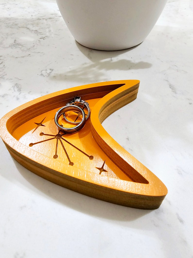 Op de afbeelding: Een halve maanvormige houten dienblad met een stervormig ontwerp en twee zilveren ringen erin. Het dienblad is in een heldere oranje kleur geverfd.