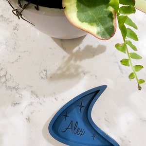 Op de afbeelding: Een blauwe keramische schaal in de vorm van een halve maan met de naam "Alex" erop. De schaal heeft aan beide kanten van de naam een stervormig ontwerp.