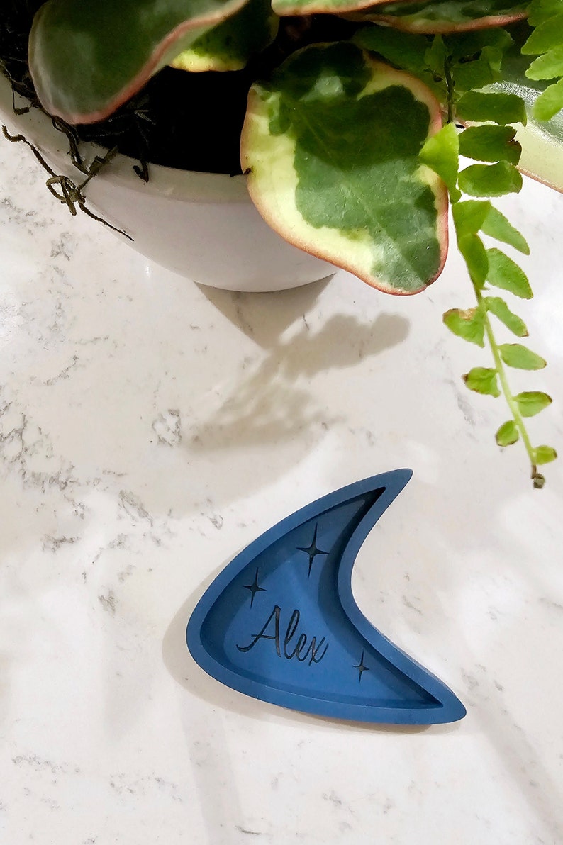 Op de afbeelding: Een blauwe keramische schaal in de vorm van een halve maan met de naam "Alex" erop. De schaal heeft aan beide kanten van de naam een stervormig ontwerp.