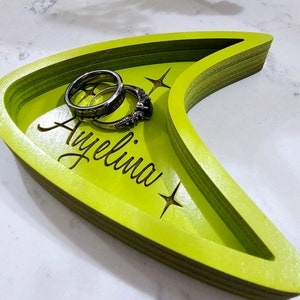 Op de afbeelding: Een groene houten dienblad in de vorm van een Star Trek-insigne met de naam "Ayelina" erop gegraveerd. Twee zilveren ringen liggen in het dienblad.