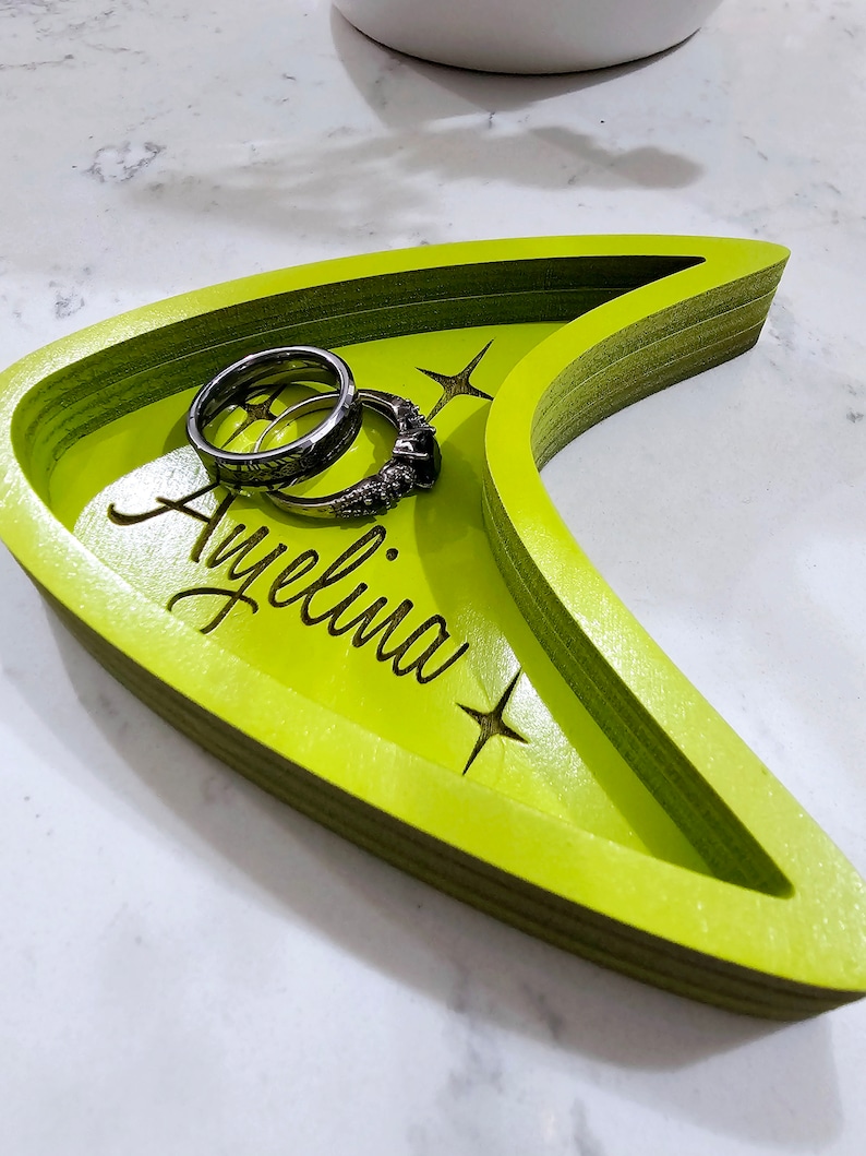 Op de afbeelding: Een groene houten dienblad in de vorm van een Star Trek-insigne met de naam "Ayelina" erop gegraveerd. Twee zilveren ringen liggen in het dienblad.