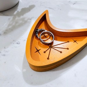 Op de afbeelding: Een houten dienblad in de vorm van een halve maan met een stervormig ontwerp en twee zilveren ringen erin. Het dienblad is in een heldere oranje kleur geverfd.