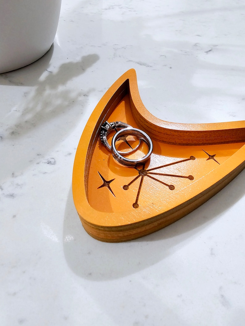 Op de afbeelding: Een houten dienblad in de vorm van een halve maan met een stervormig ontwerp en twee zilveren ringen erin. Het dienblad is in een heldere oranje kleur geverfd.