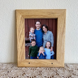 Puede incluir: Una fotografía enmarcada de una familia de cinco personas. La familia está de pie frente a una puerta de madera roja. Los padres sostienen a dos de los niños, mientras que el niño más pequeño está de pie frente a ellos. La foto está en un marco de madera marrón claro.