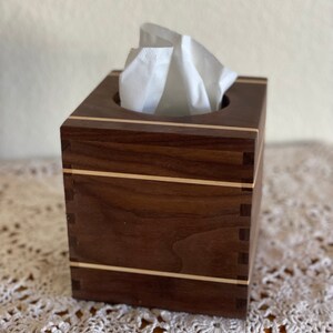May include: A square wooden tissue box with a dark brown finish and light wood accents. The box is filled with white tissues, and the construction features visible dovetail joints. The design is simple and elegant.