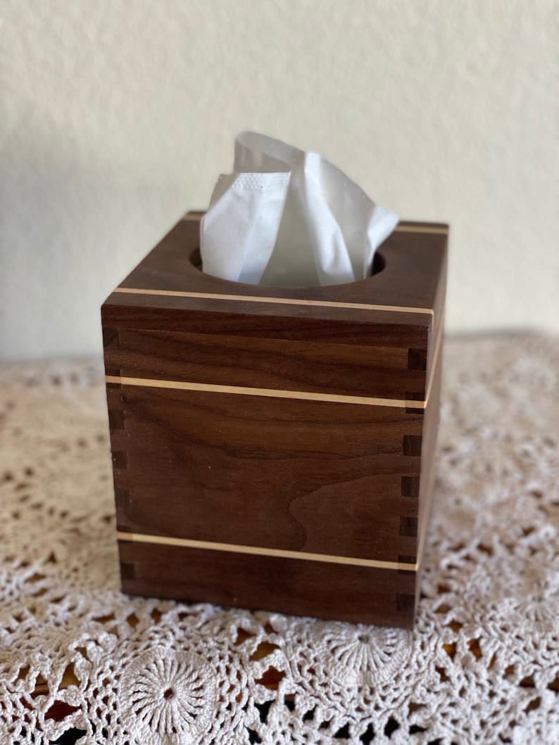 May include: A square wooden tissue box with a dark brown finish and light wood accents. The box is filled with white tissues, and the construction features visible dovetail joints. The design is simple and elegant.