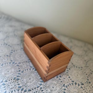 May include: A wooden desk organizer with three compartments. The organizer is made of light brown wood with visible dovetail joints and a curved top. The compartments are open, and the organizer sits on a white and blue patterned surface.