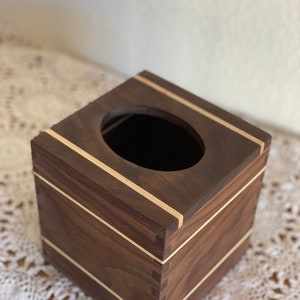 May include: A square wooden tissue box with a dark brown finish and light-colored accent stripes. The box has a circular opening on top for tissue dispensing. The corners of the box are joined with dovetail joints.